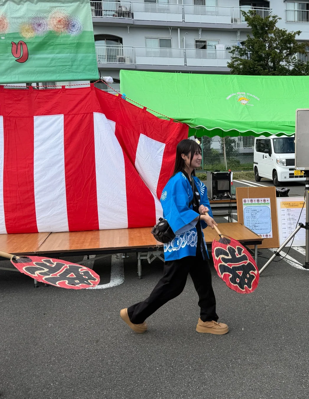 青い法被を着た女性が「祭」の文字が入ったうちわを持ち、日本の祭りの会場を歩いている様子