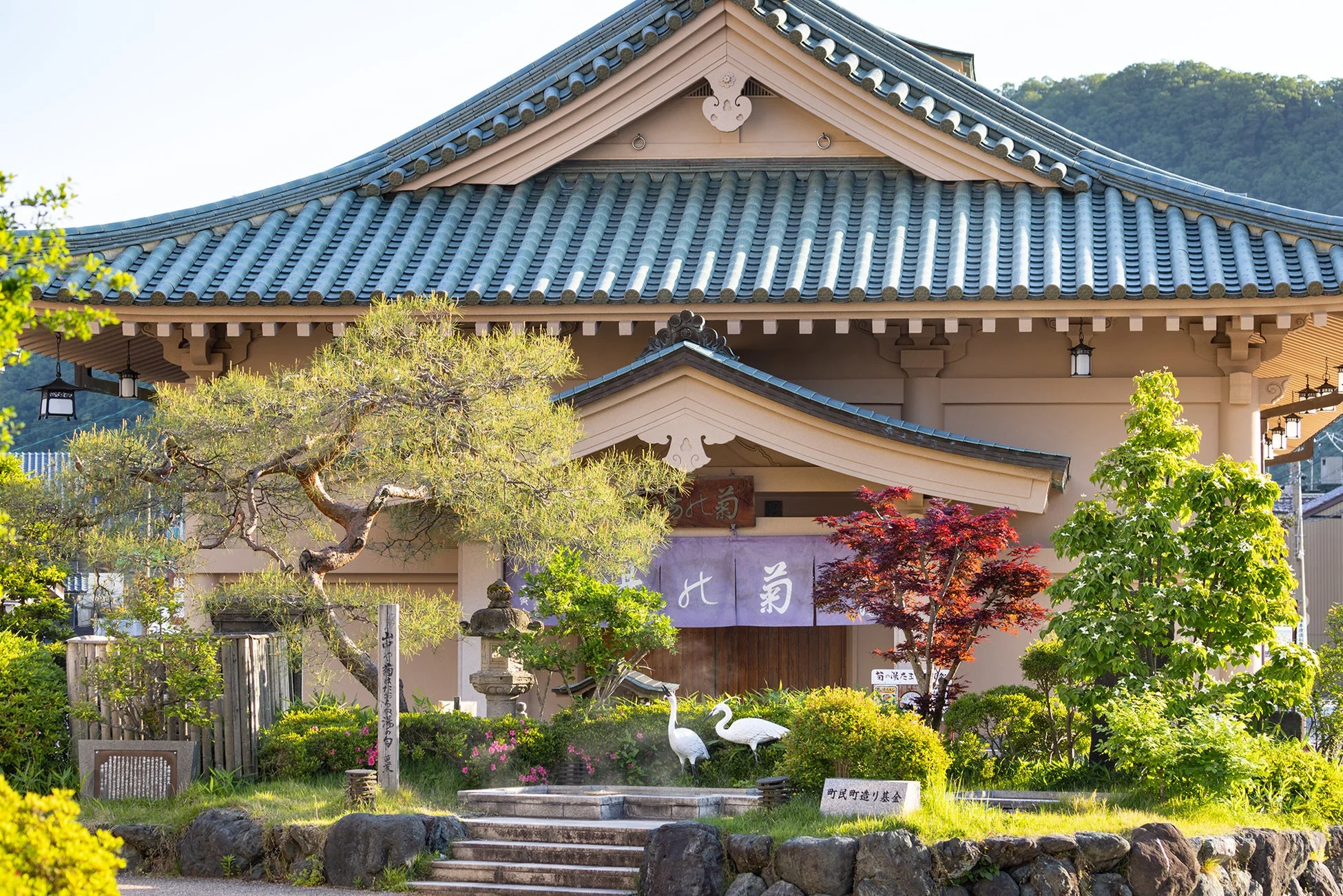 「鶴の湯」という伝統的な日本建築の温泉施設を捉えた一枚。青い瓦屋根が印象的で、手入れの行き届いた日本庭園には石灯籠や鶴の像が配置され、豊かな自然に囲まれた趣のある風景が広がっています。
