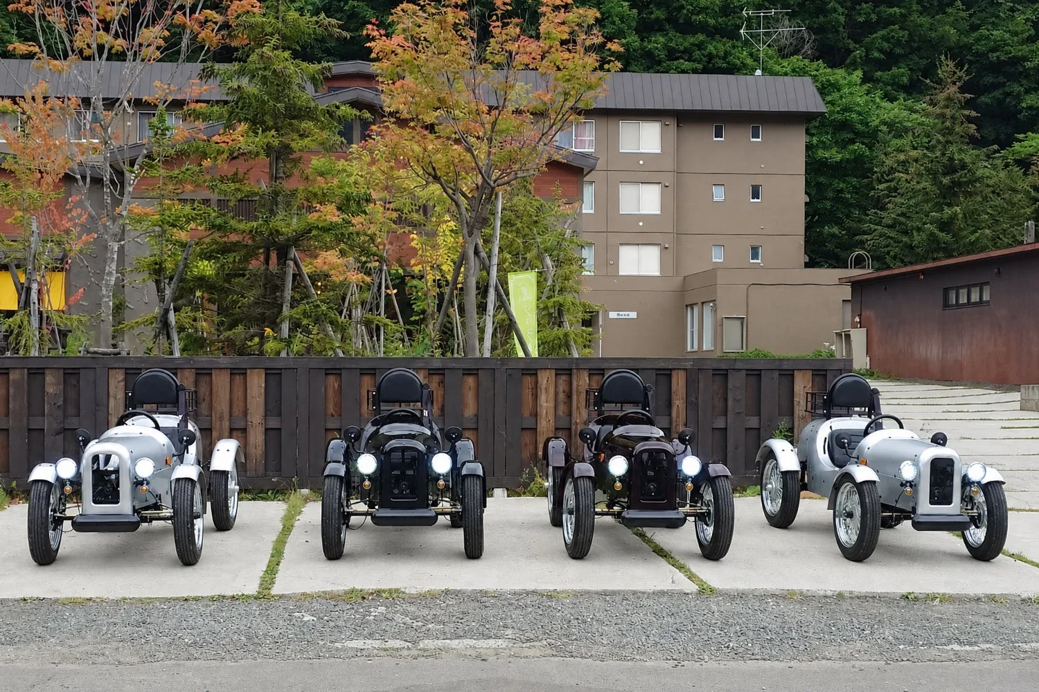 秋の紅葉が美しい背景に、レトロなデザインの小型車が4台並んで駐車されています。