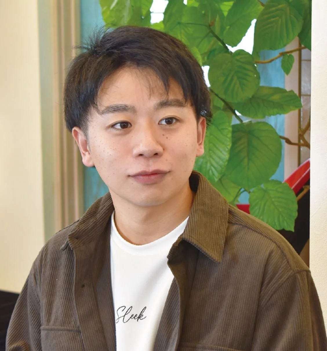 茶色のコーデュロイシャツと白いTシャツを着た若いアジア人男性のポートレートです。男性は少し右方向を見ており、背景には緑の葉の植物が見えます。