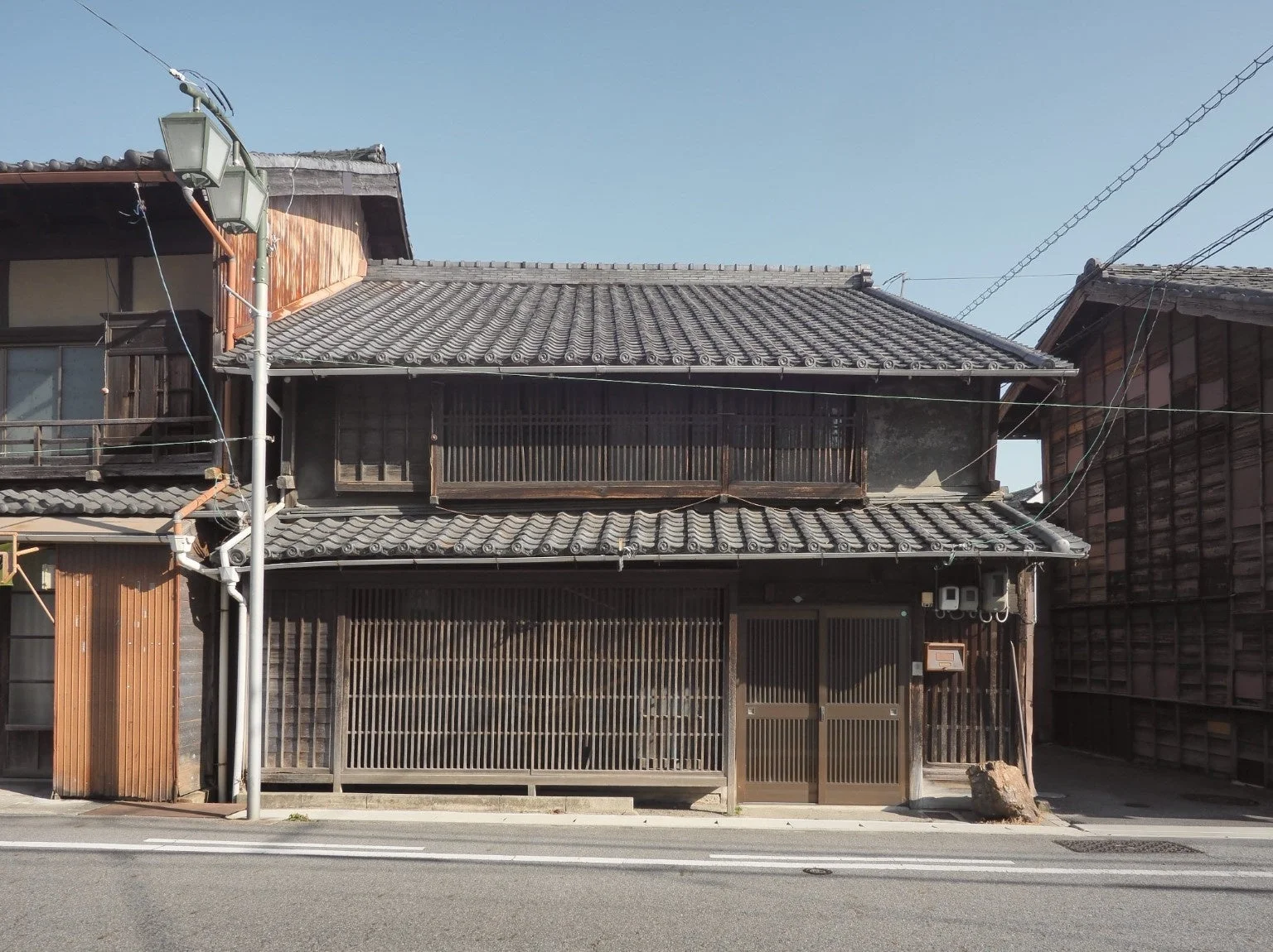 瓦屋根と格子戸が特徴的な日本の伝統的な町家が建ち並ぶ静かな街並み。手前には電柱と街灯があり、穏やかな日中の風景が広がっています。