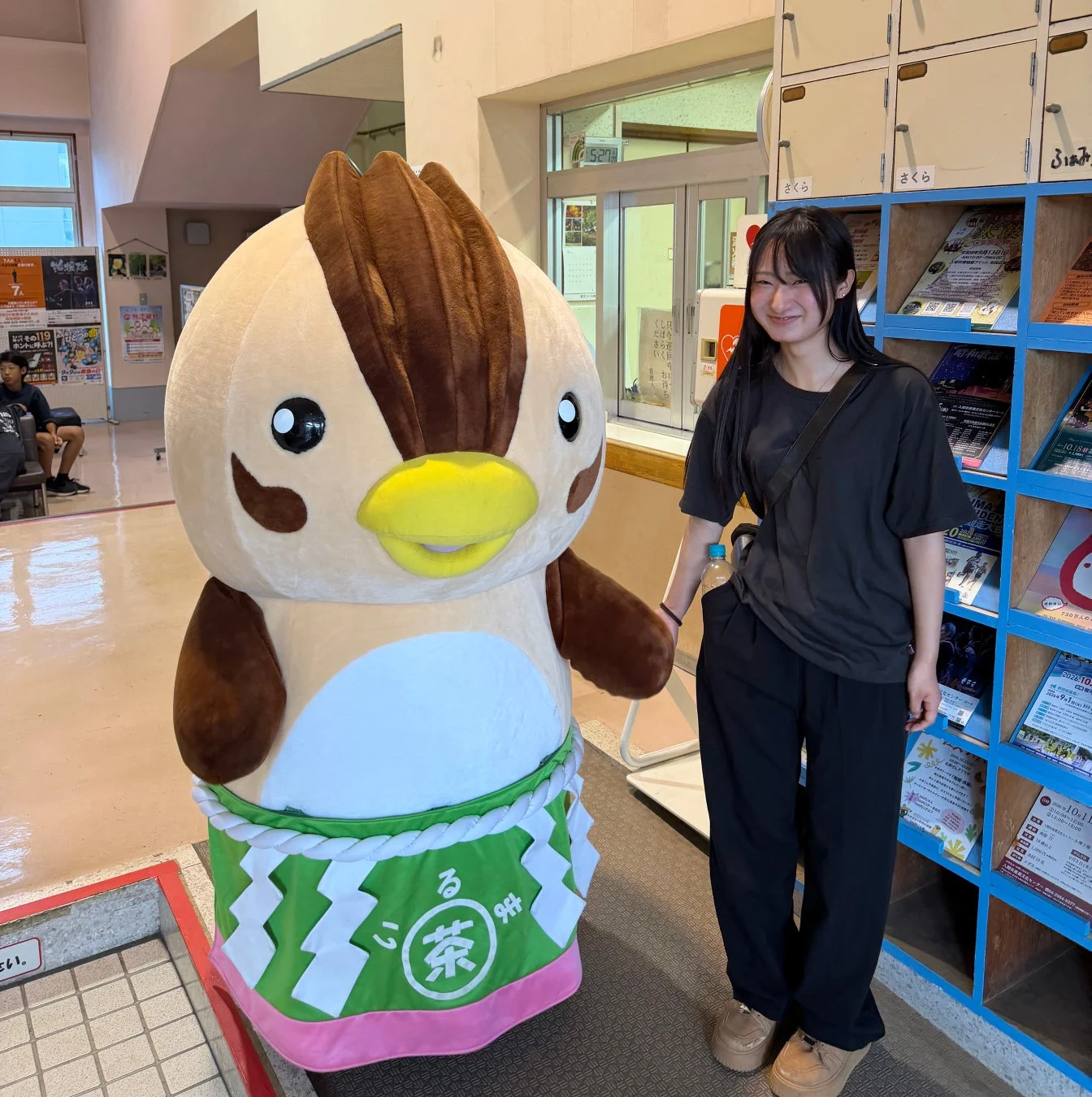 可愛らしい鳥のゆるキャラと黒いTシャツを着た若い女性が屋内で並んで立っている