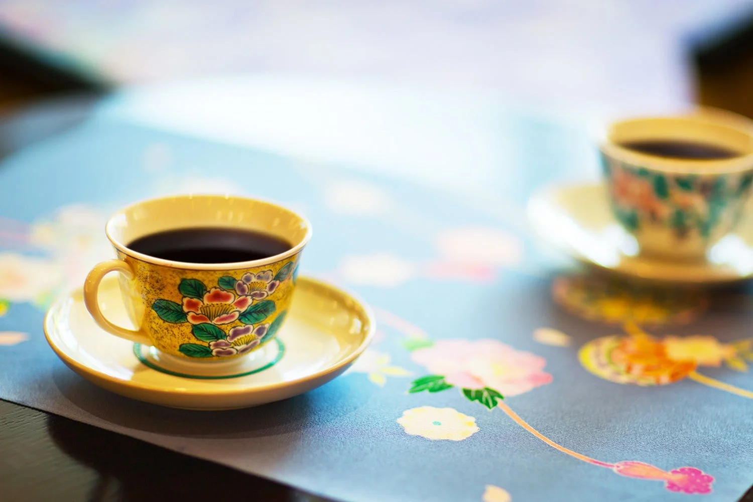 美しい花柄のカップ＆ソーサーに入ったコーヒーが、花模様のランチョンマットの上に置かれています。手前のカップが鮮明で、優雅なひとときを感じさせます。