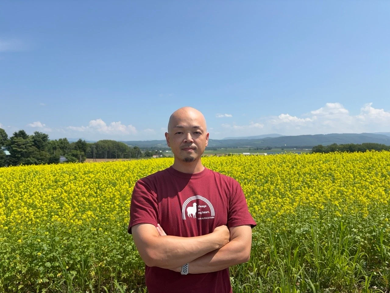 晴れた青空の下、一面に広がる黄色い菜の花畑を背景に、坊主頭の男性が腕を組んで立っています