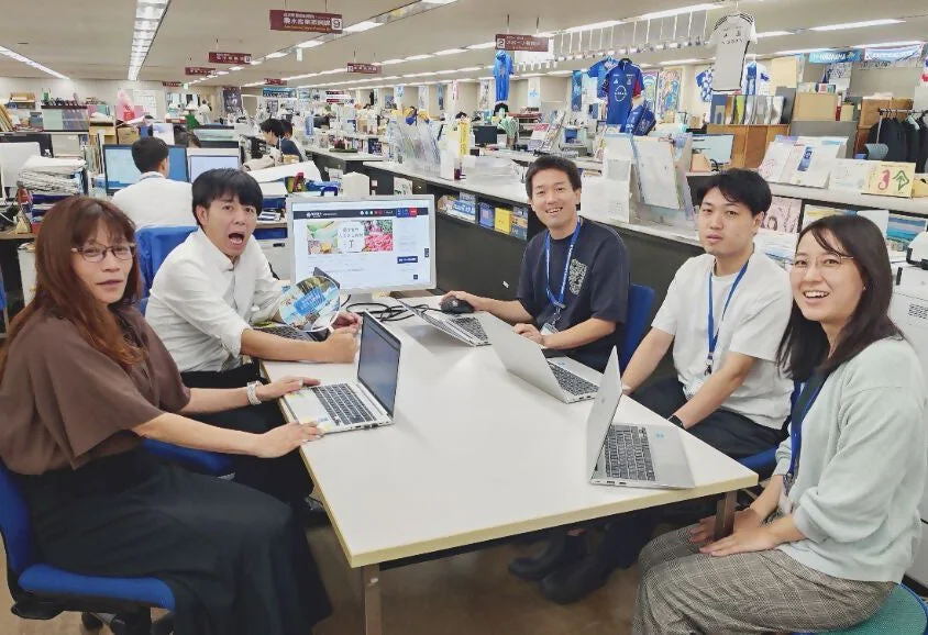 オフィス内で5人の男女が机を囲み、ノートパソコンやデスクトップPCを使って作業している様子。皆が笑顔を見せる中、一人だけ驚いた表情をしており、和やかながらも活気のあるチームの雰囲気が伝わってきます。