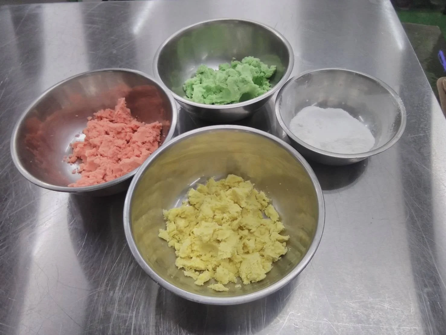お菓子作りの準備風景。ステンレス製のボウルにピンク、緑、黄色のカラフルな生地と白い粉が入っている