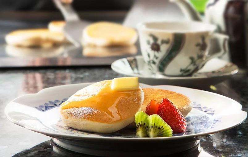 ふわふわのパンケーキにバターとシロップがたっぷりかかっており、いちごとキウイが添えられています。奥にはティーカップが見え、温かい飲み物と一緒に楽しめる美味しそうなデザートの光景です。