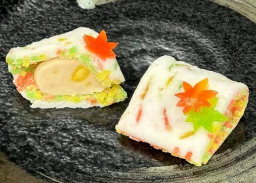 黒い皿に盛られた、秋の紅葉をモチーフにした美しい和菓子が2つ。白い外皮の中には黄色い餡が見える