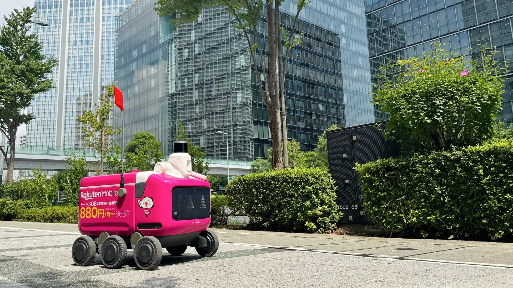楽天モバイルのロゴが入ったピンク色の自動配送ロボットが、高層ビルが立ち並ぶ都市の歩道を進んでいます。