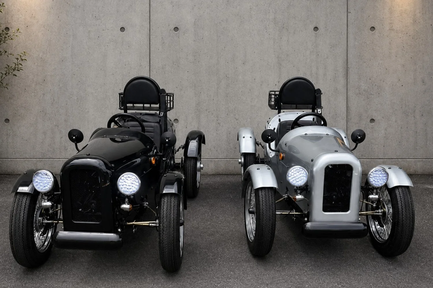 コンクリートの壁を背景に、黒とシルバーのレトロなデザインの三輪自動車が2台並んで駐車している様子。
