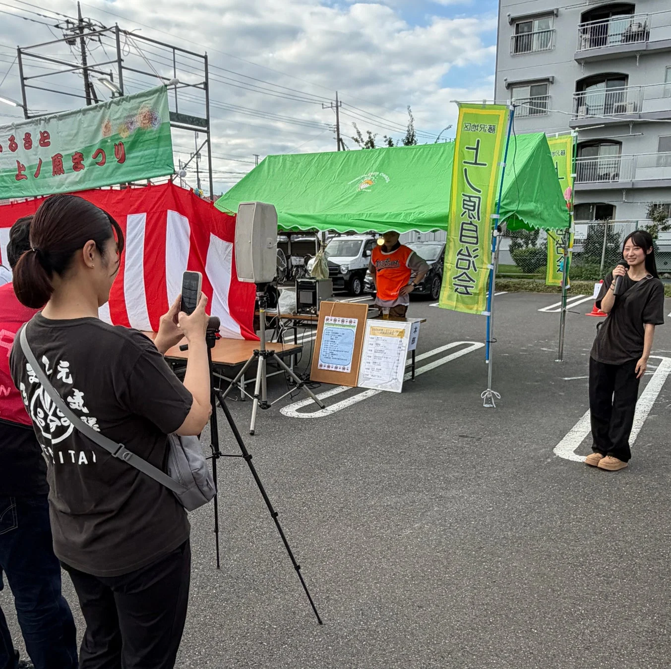 屋外の地域イベントで、女性がマイクを持って話しており、別の女性がスマートフォンで撮影している様子
