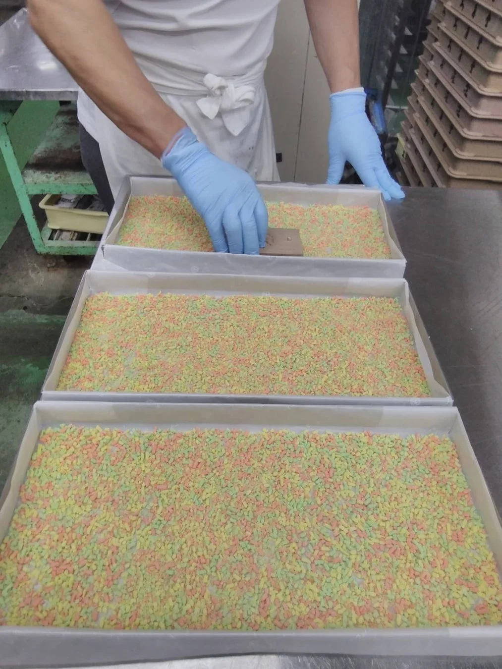 白いエプロンと青い手袋を着用した人物が、カラフルな小粒の菓子が敷き詰められた複数のトレーの上で作業している