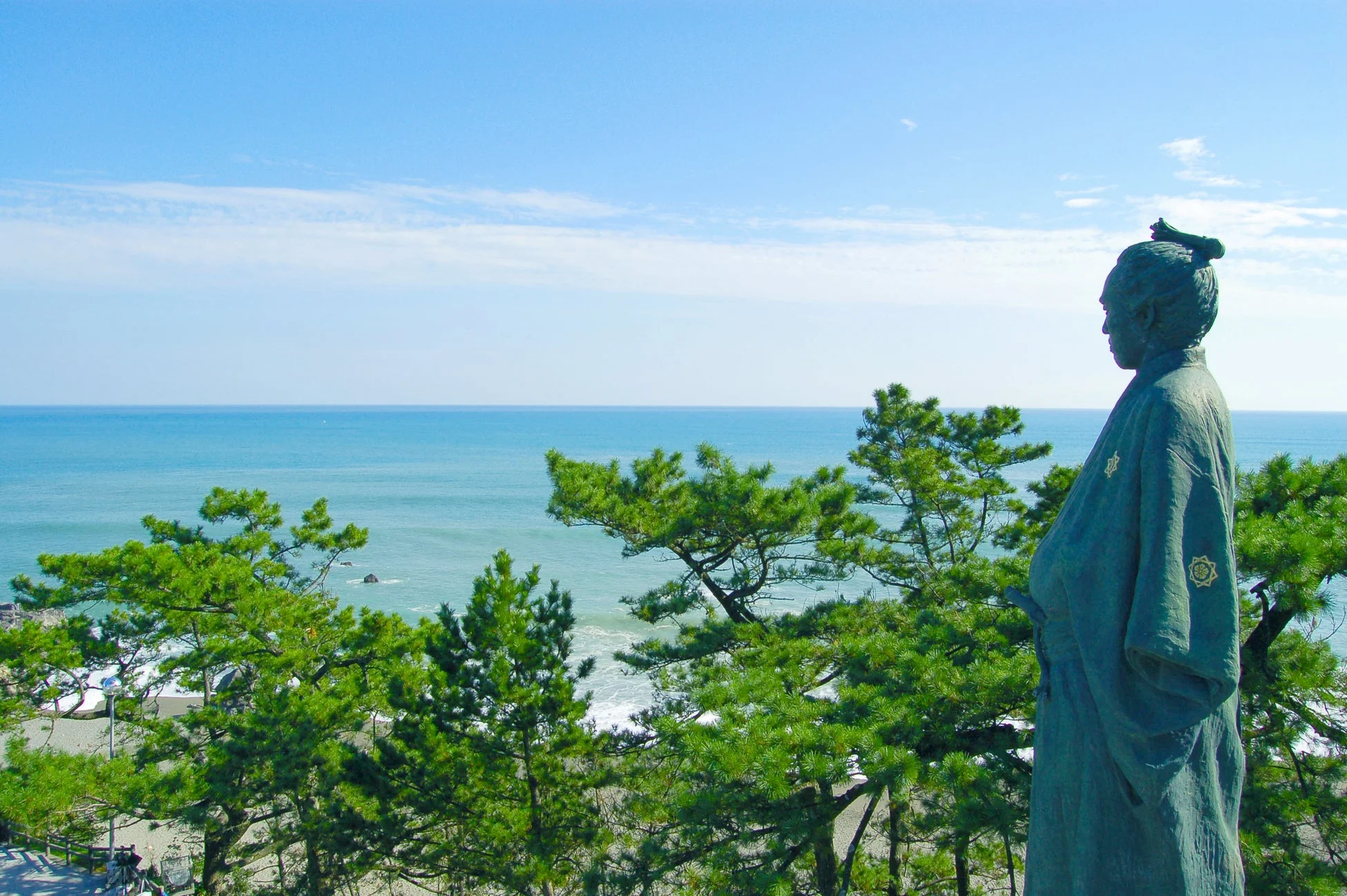 青い海と空を背景に、松の木々に囲まれた歴史的な人物の銅像が佇む風景。穏やかな海岸線の景色が広がっています。